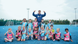 A group of children wearing soccer bibs pose with a coach on a blue outdoor soccer field.