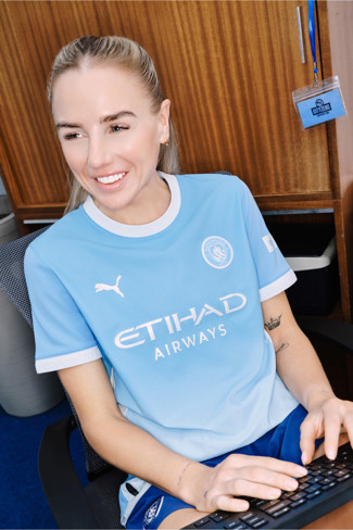 A person with their face blurred is wearing a Manchester City jersey while typing on a keyboard. The setting appears to be an office with a wooden cabinet and a lanyard badge nearby.