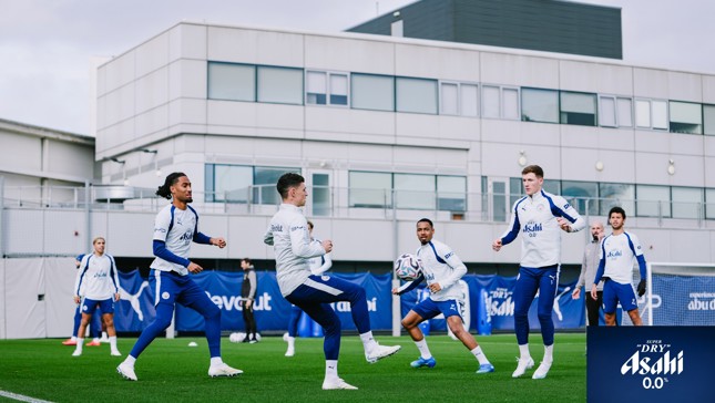 Manchester City players engaged in a training session on a grassy field wearing branded training gear, surrounded by buildings.