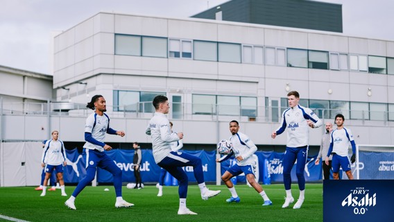 Manchester City players engaged in a training session on a grassy field wearing branded training gear, surrounded by buildings.