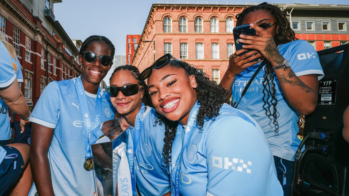 A group of people in Manchester City jerseys celebrating with medals around their necks in an outdoor urban setting.