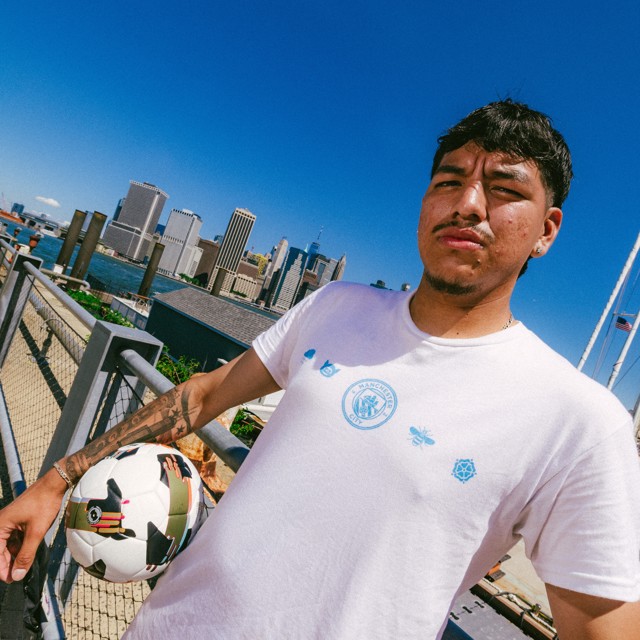 Person holding a soccer ball wearing a Manchester City t-shirt with New York skyline in the background.