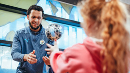 A Manchester City staff member wearing a jacket with the team logo interacts with a young fan tossing a ball indoors.