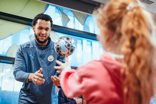 A Manchester City staff member wearing a jacket with the team logo interacts with a young fan tossing a ball indoors.