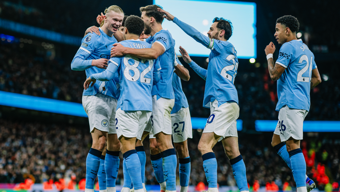 A group of Manchester City players celebrating on the field in light blue jerseys with sponsor logos. The number 82 is visible on one player’s back.