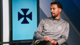 A person in a gray Manchester City tracksuit featuring the Puma logo and initials 'LC' is seated near a screen displaying a black symbol.
