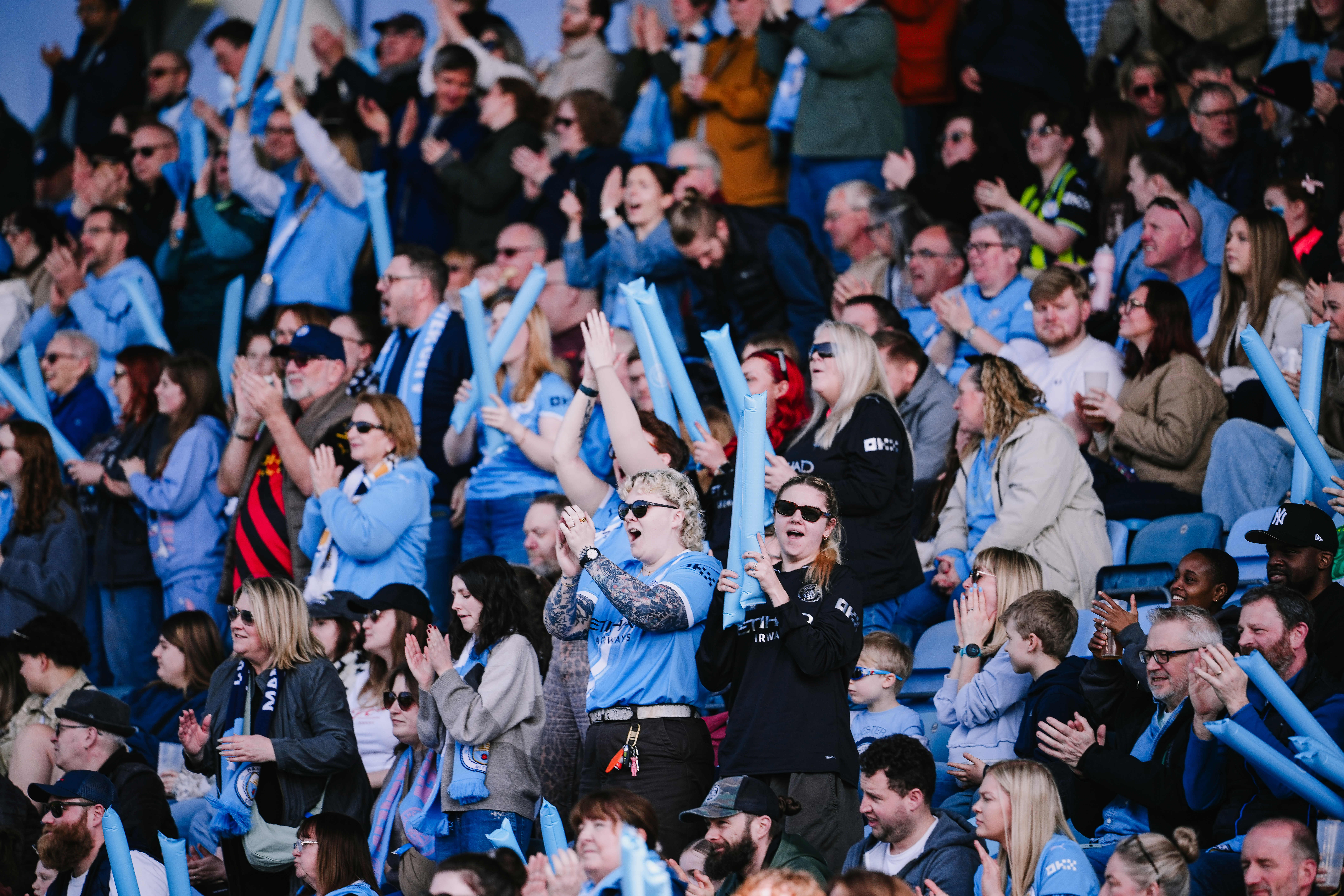 A crowd of enthusiastic fans wearing light blue clothing, cheering and raising inflatable sticks at a sporting event.