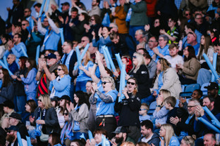 A crowd of enthusiastic fans wearing light blue clothing, cheering and raising inflatable sticks at a sporting event.