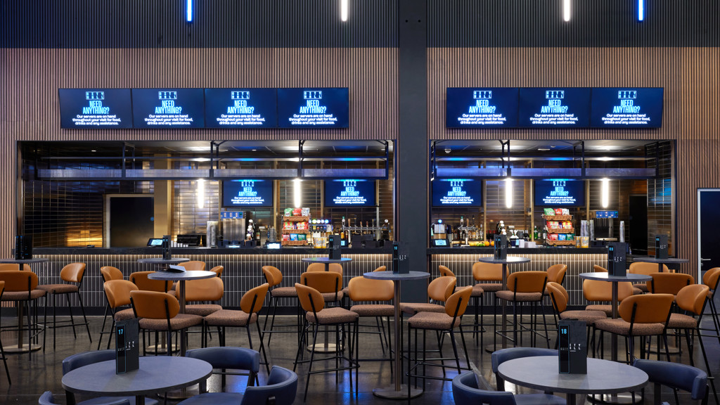 Modern bar and lounge area with seating, tables, and digital screens displaying services available.