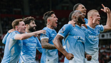 A group of Manchester City players wearing light blue jerseys are celebrating together on the field, displaying expressions of joy and excitement.