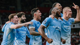 A group of Manchester City players wearing light blue jerseys are celebrating together on the field, displaying expressions of joy and excitement.