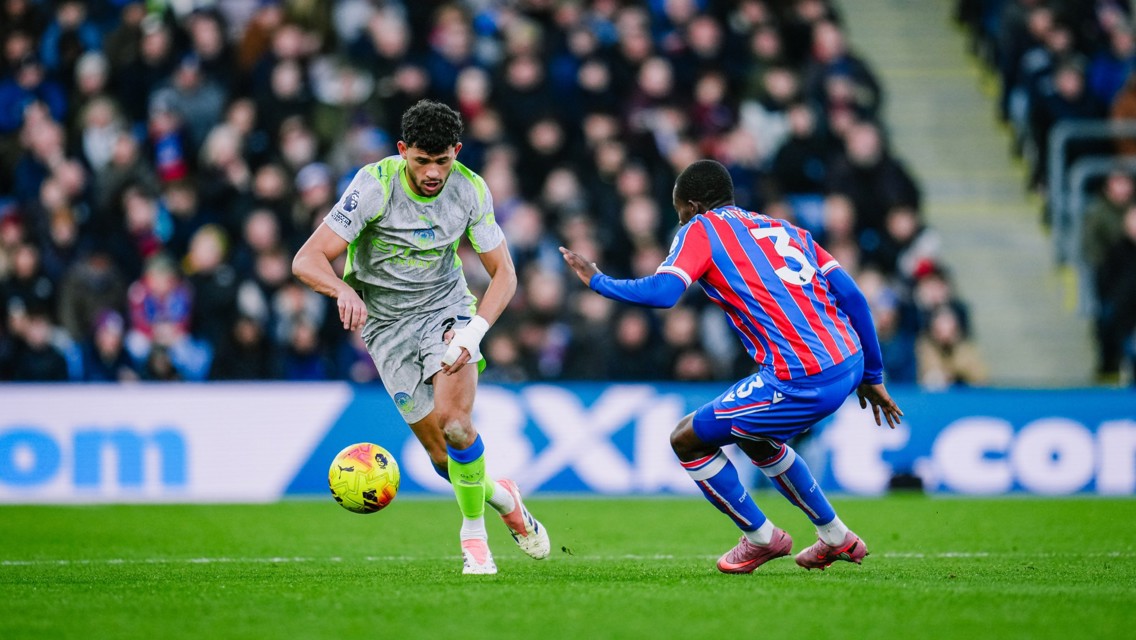 Two football players from opposing teams in action during a match, with one player in a gray and neon kit running with the ball and the other in a red and blue striped kit defending. The crowd watches in the background.