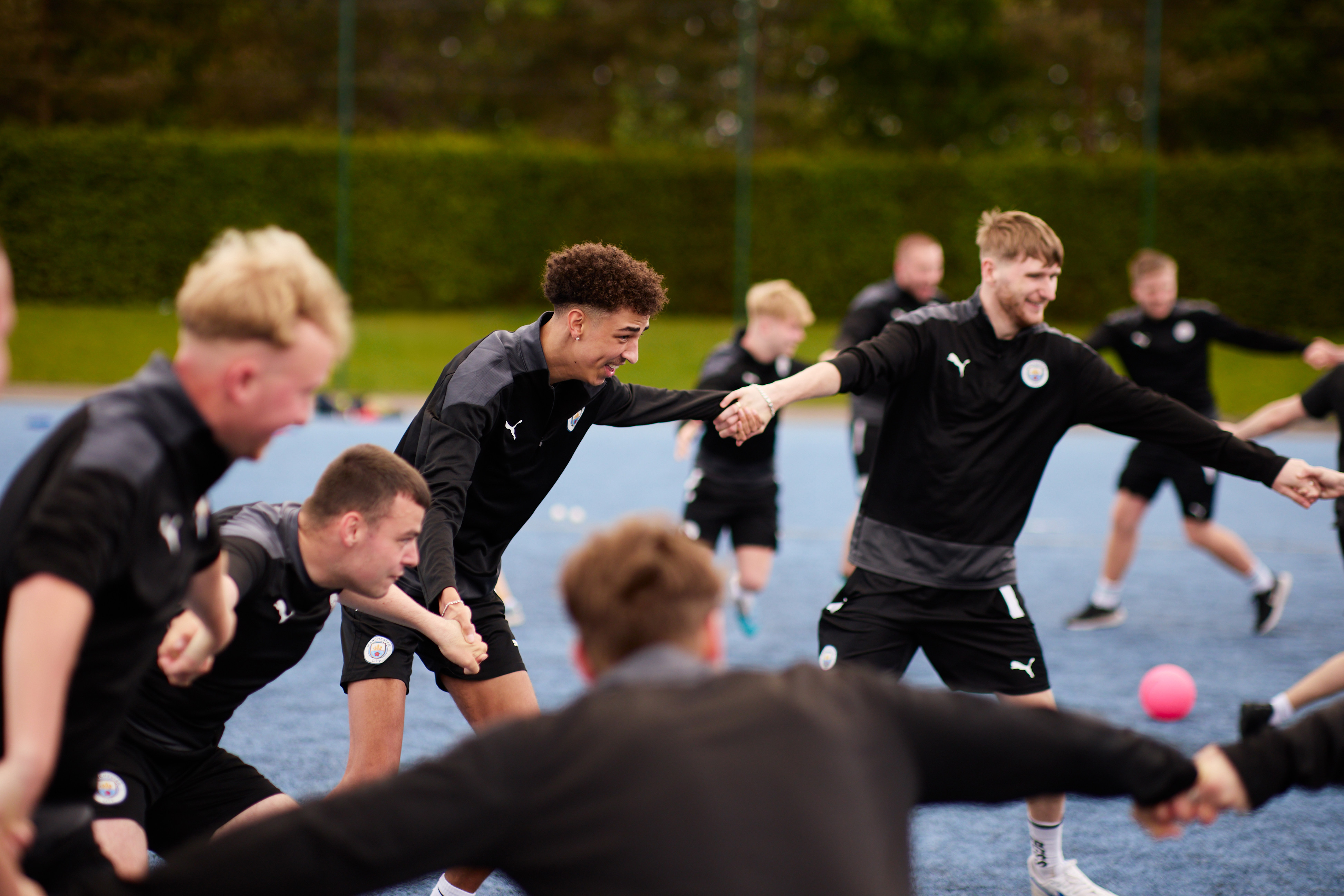 Players of Manchester City perform stretching exercises during a training session on a blue pitch.