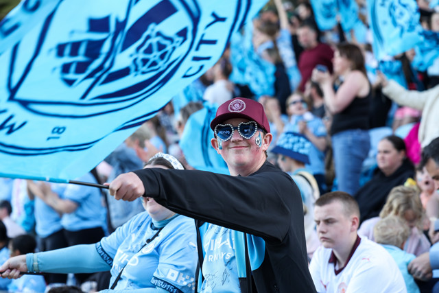 Fans of Manchester City, wearing light blue jerseys, waving flags with the Manchester City crest at a stadium event.