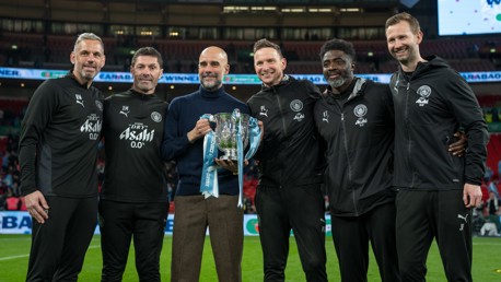 A group of six people in Manchester City attire holding a trophy with ribbons on a football field.