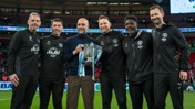 A group of six people in Manchester City attire holding a trophy with ribbons on a football field.