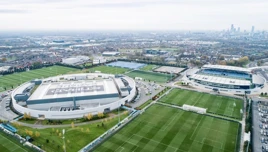 Aerial view of Manchester City's Etihad Campus with multiple football pitches, the City Football Academy, and the Academy Stadium. The city skyline is visible in the background.