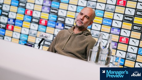 A person with a blurred face in a press conference setting featuring Manchester City logos and sponsors on the backdrop.