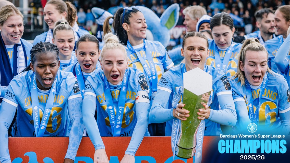 Team photo of Manchester City Women's team celebrating as Barclays Women's Super League Champions for the 2025/26 season. Players are in light blue kits, wearing gold 'Champions' scarves and holding the trophy.