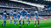 Manchester City players celebrating on the field at Etihad Stadium, wearing light blue kits with a crowded stadium in the background.