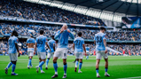 Manchester City players celebrating on the field at Etihad Stadium, wearing light blue kits with a crowded stadium in the background.