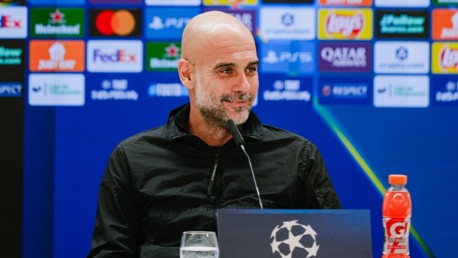 A person is speaking at a press conference, the background features Champions League sponsor logos, and a Gatorade bottle is visible on the table.