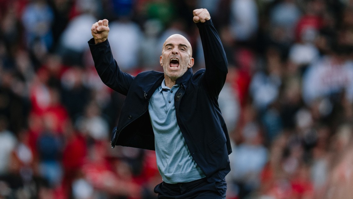 A person in a black jacket and grey shirt celebrates with clenched fists in a crowded sports stadium.