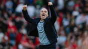 A person in a black jacket and grey shirt celebrates with clenched fists in a crowded sports stadium.