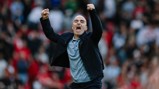 A person in a black jacket and grey shirt celebrates with clenched fists in a crowded sports stadium.