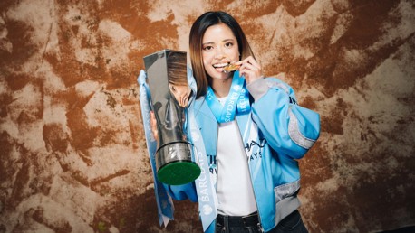 Person holding a large trophy and biting a medal, wearing a blue jacket with a Barclays ribbon.