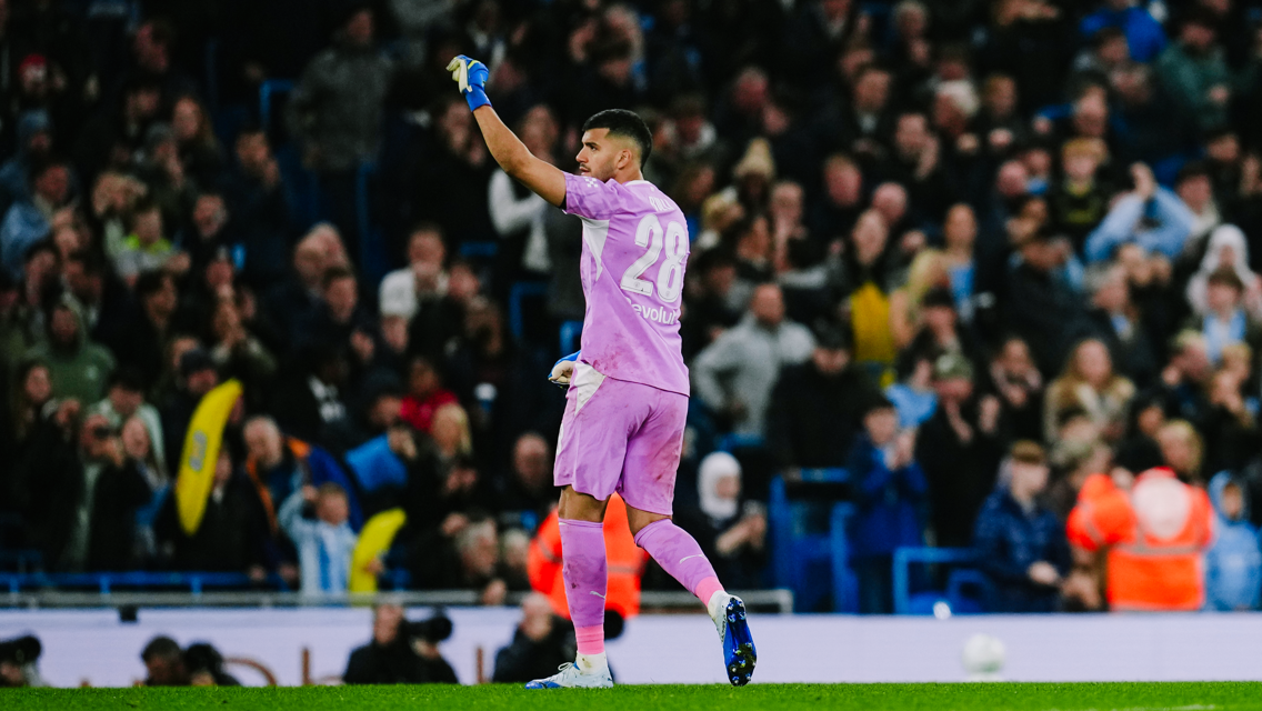 A goalkeeper wearing a pink uniform with the number 28, raising a hand, during a match with a blurred crowd in the background.