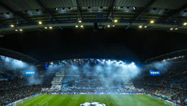 Night football match at Etihad Stadium with fog and player line-up in the center. Stadium lights illuminate the scene, and fans fill the stands with blue and white displays.