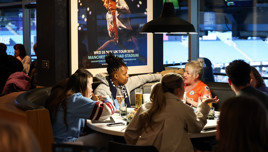 A group of people sitting at a round table in a restaurant, engaged in conversation. One person is wearing a Manchester City jersey, and a poster for a concert at Etihad Stadium is visible.