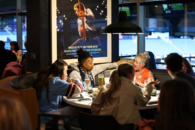 A group of people sitting at a round table in a restaurant, engaged in conversation. One person is wearing a Manchester City jersey, and a poster for a concert at Etihad Stadium is visible.