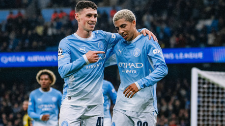 Two Manchester City players in light blue jerseys celebrate a goal during a match, with one player pointing at the other.