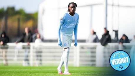 A Manchester City youth player in a blue kit stands on a soccer field during a match. The 'Official Verdict' logo is visible.