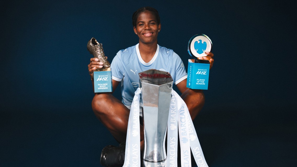 A soccer player holding Barclays WSL Golden Boot and Player of the Season trophies, Manchester City jersey, with a silver trophy in front.