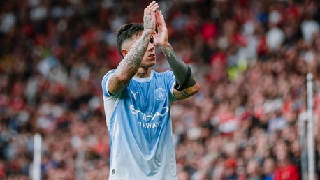 A soccer player in a Manchester City uniform clapping on the field, with visible tattoos on arms. The crowd is blurred in the background.