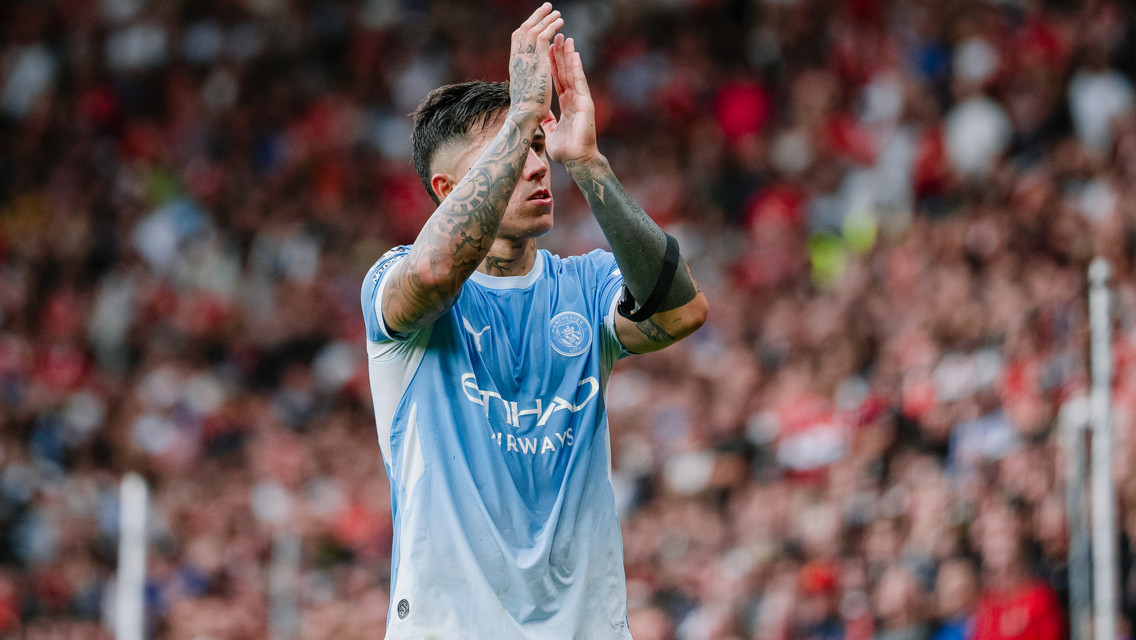 A soccer player in a Manchester City uniform clapping on the field, with visible tattoos on arms. The crowd is blurred in the background.