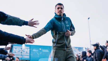 A person wearing a Manchester City tracksuit high-fives fans in front of a team bus. The Manchester City logo is visible on the clothing.