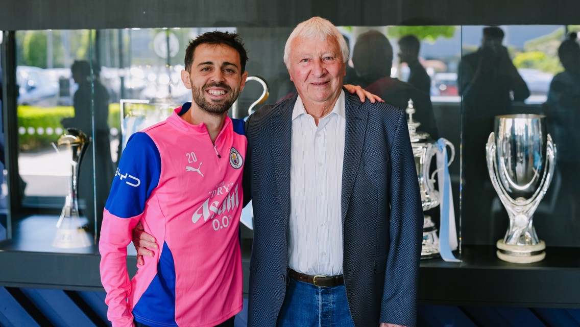 Two people, one in a Manchester City jersey, pose in a trophy room with multiple trophies in the background.