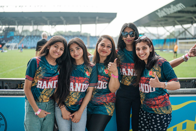 Five women wearing green and red Bandung shirts posing together with a stadium in the backdrop.
