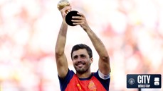 A football player in a red and blue kit holds a golden trophy above his head, smiling. The background is blurred, emphasizing his gesture of victory.
