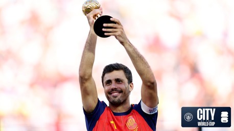 A football player in a red and blue kit holds a golden trophy above his head, smiling. The background is blurred, emphasizing his gesture of victory.