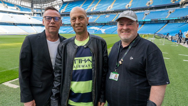 Three men posing for a photo on the pitch of the Etihad Stadium, home of Manchester City Football Club.