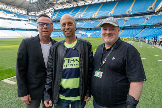 Three men posing for a photo on the pitch of the Etihad Stadium, home of Manchester City Football Club.