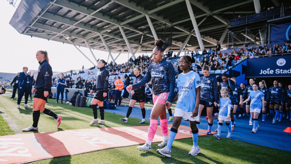 Players and children walk out on the field at Joie Stadium, home of Manchester City, with the crowd in the background.