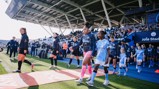 Players and children walk out on the field at Joie Stadium, home of Manchester City, with the crowd in the background.