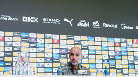 A press conference background with sponsorship logos and a blurred face at the front table.