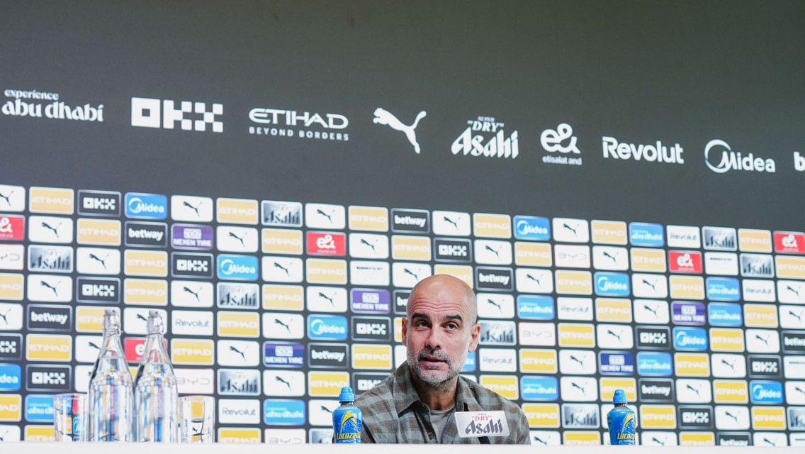 A press conference background with sponsorship logos and a blurred face at the front table.